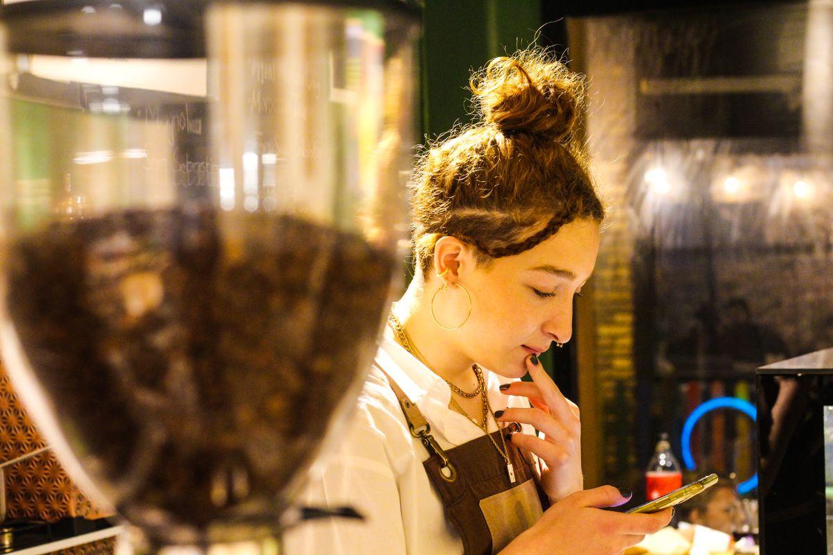 A focused young barista checks her phone while working in a cozy coffee shop