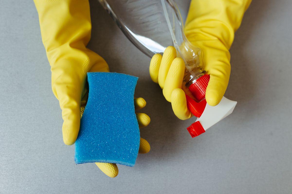 Hands wearing yellow gloves holding a sponge and a spray bottle for cleaning