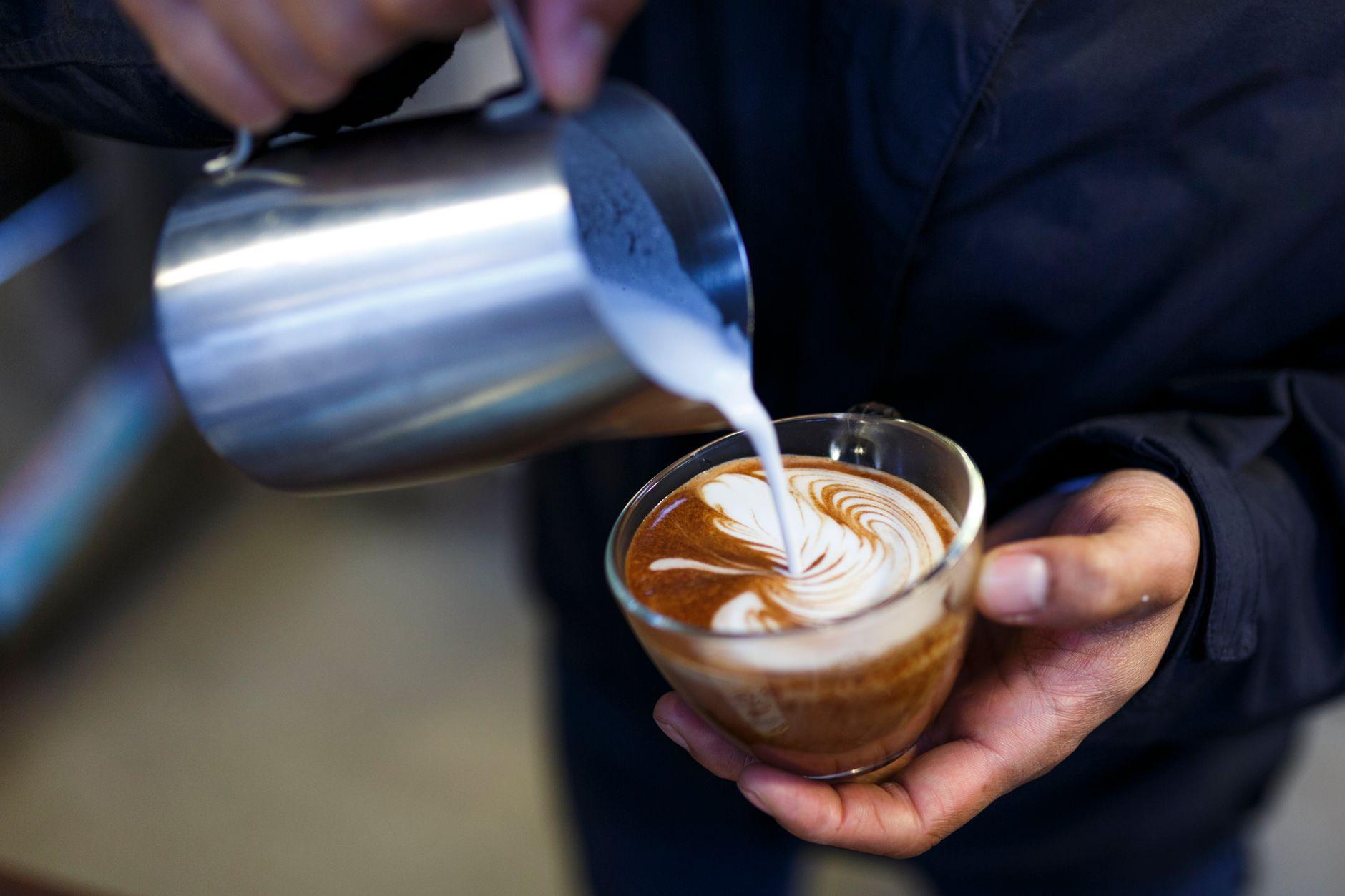 Posnetek od blizu: baristka prelije soparo mleko in ustvara zapleten latte art v stekleni skodelici