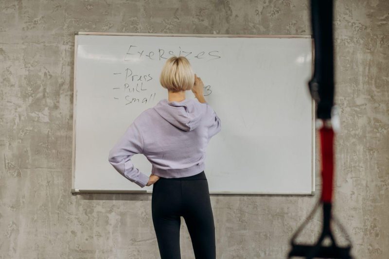 Female fitness instructor writing exercises on a whiteboard in a gym, planning the weekly workout schedule