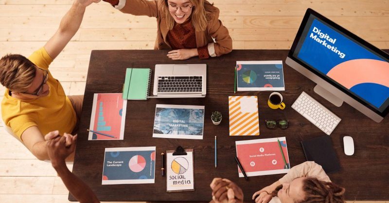 Marketing team collaborating around a desk in a modern office with laptops and screens showing social media analytics