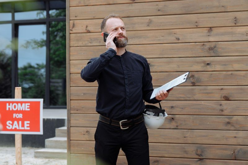 Real estate agent making a phone call while holding documents near a home for sale sign