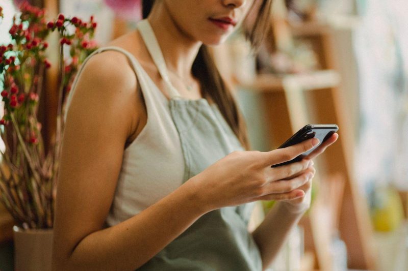 Florist in apron browsing mobile phone while working in cozy floral shop