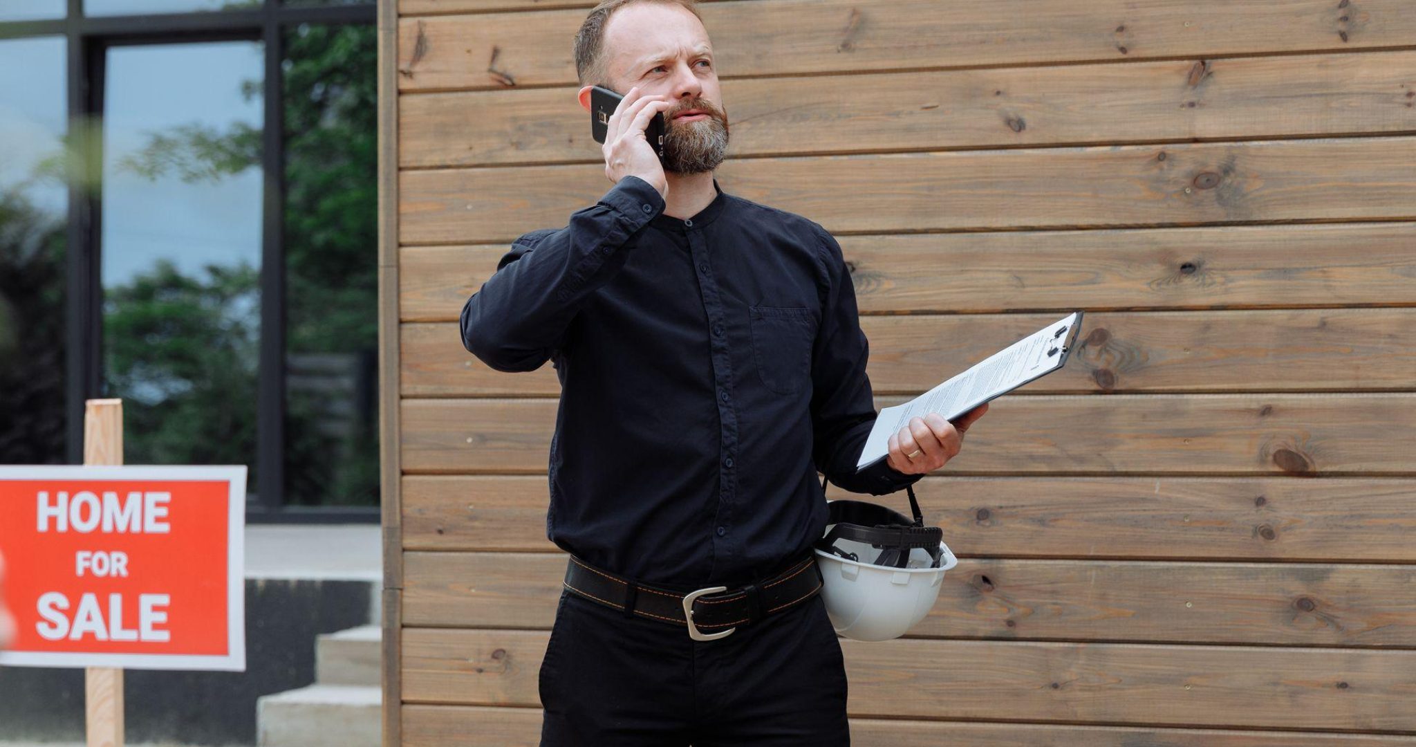 Real estate agent making a phone call while holding documents near a home for sale sign